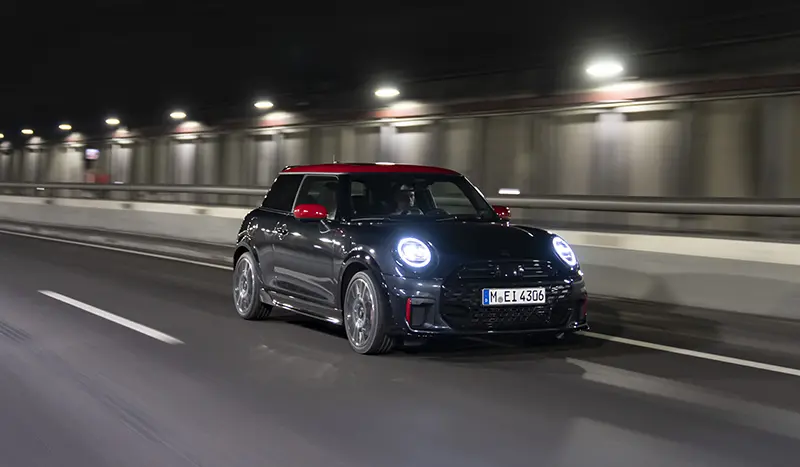 Mini John Cooper Works Hatchback driving through a lit tunnel, showcasing its illuminated headlights.