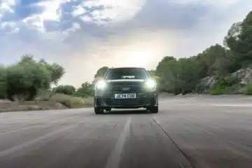 Front view of the new Mini John Cooper Works Hatchback driving on a scenic road with headlights on.