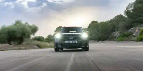 Front view of the new Mini John Cooper Works Hatchback driving on a scenic road with headlights on.