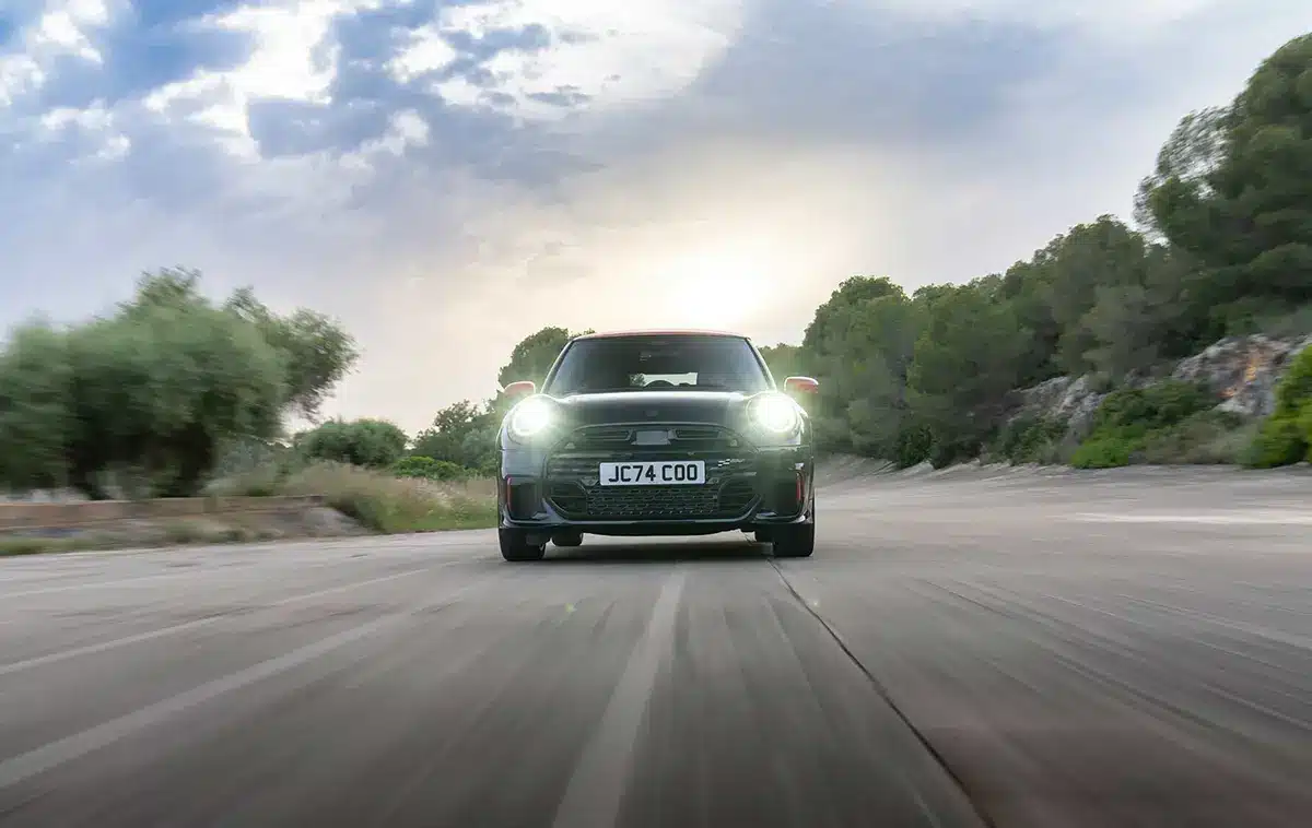 Front view of the new Mini John Cooper Works Hatchback driving on a scenic road with headlights on.