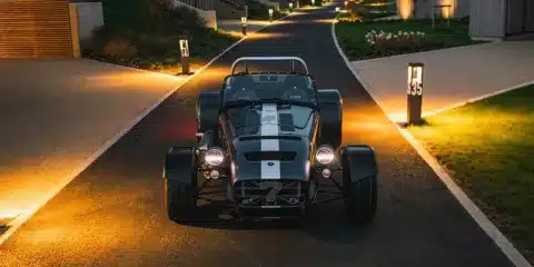 Caterham Seven CSR Twenty driving at sunset with headlights illuminating a warm-lit road.