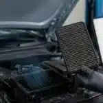 Mechanic comparing a clogged engine air filter with a clean replacement beside an open car engine air filter housing