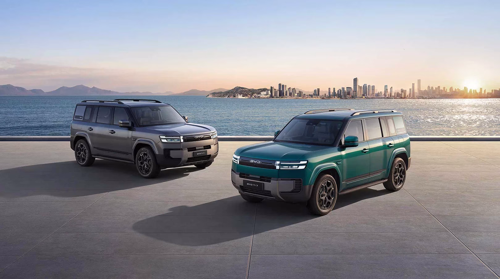 Green and grey BYD Ti7 SUVs parked beside the water with a city skyline behind