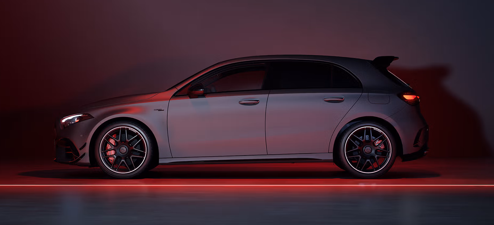 Silver-grey Mercedes-AMG A45 S in full side profile in a dark studio with red lighting, showing its roof spoiler and matte black AMG forged wheels