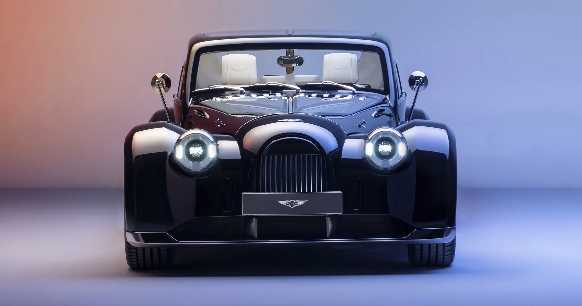 Dark blue Morgan Midsummer Coupé head-on in a studio, round headlights lit, showing its classic Morgan grille and glass roof