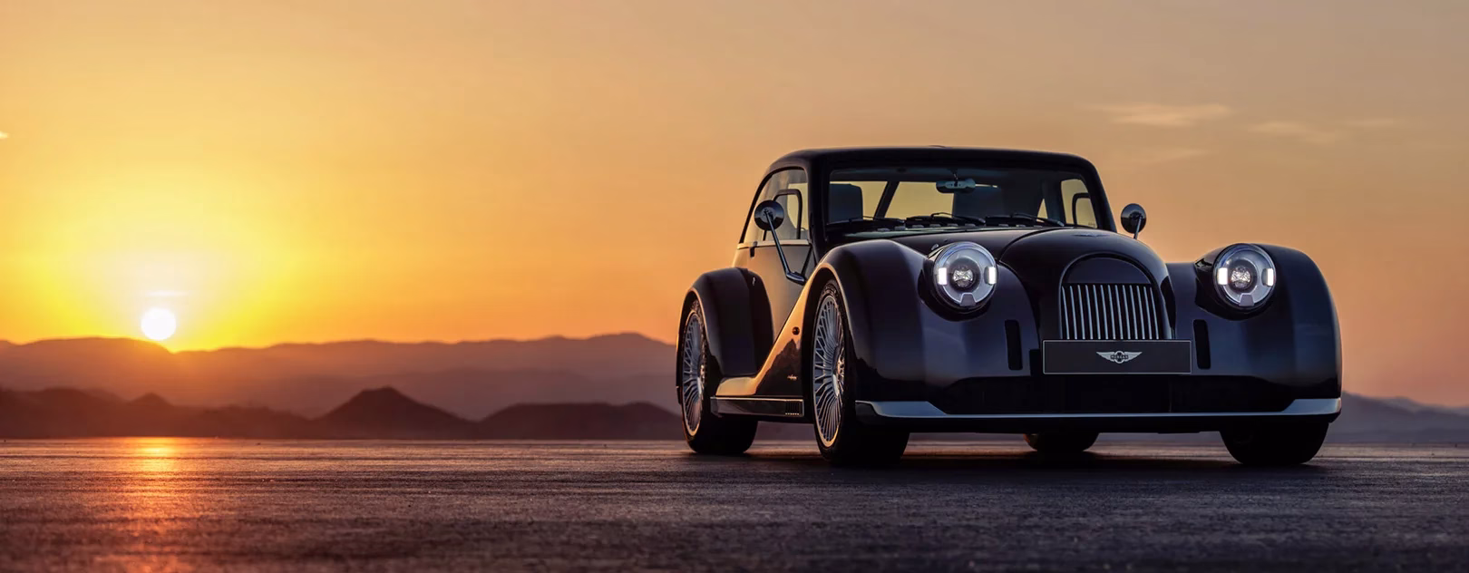 Dark Morgan Midsummer Coupé parked on an open plain at sunset, front three-quarter, sun low on the horizon