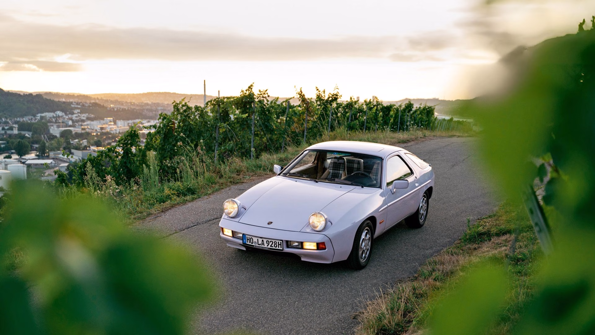 Pale lilac Porsche 928 on a vineyard road at sunset with a city below